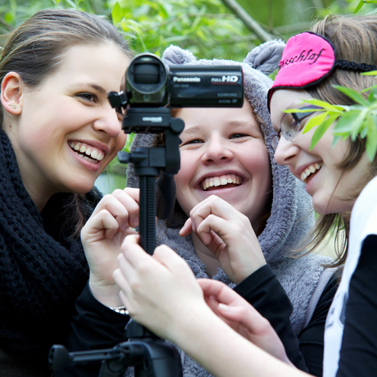 Drei Mädchen sind verkleidet und schauen lachend in eine Filmkamera