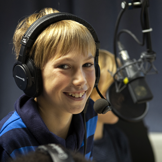 Junge mit Kopfhörern sitzt vor einem Studiomikrofon und lacht ins Bild