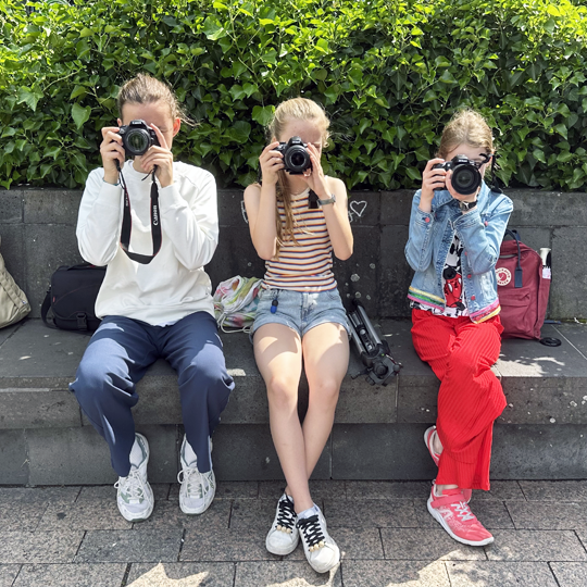 Drei Kinder sitzen auf einer Bank und schauen durch ihre Fotokamera ins Bild
