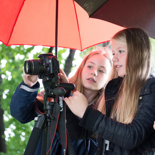 Zwei Mädchen unter Regenschirmen schauen gemeinsam durch eine Fotokamera